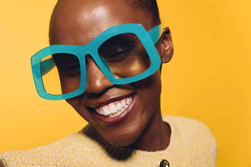 Smiling dark skinned African woman wearing oversized blue glasses and yellow sweater against bright yellow background. Concept of fashion, joy, beauty, and vibrant lifestyle.