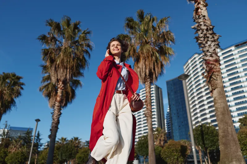 Confident woman in a red coat walks through a sunny urban park, palm trees and modern buildings in the background, creating a cheerful city lifestyle moment.