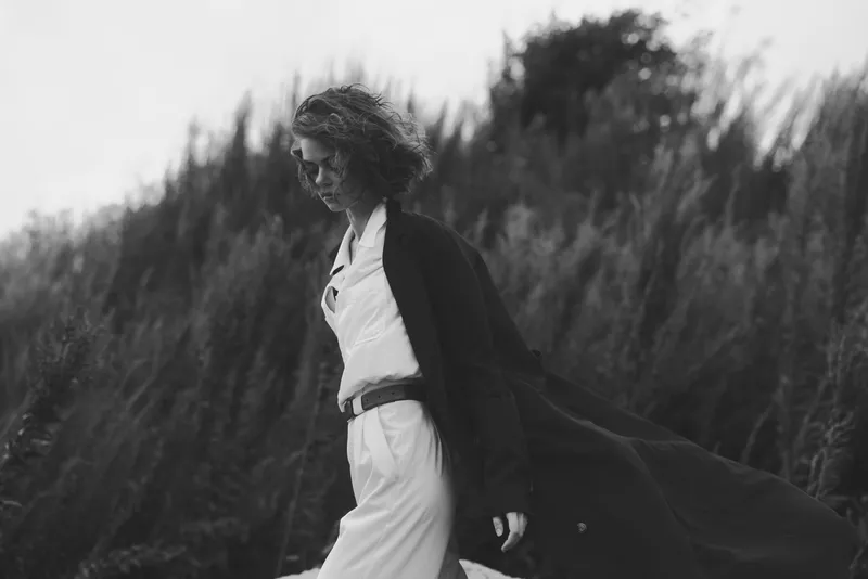 Woman wearing a flowing cape walks along a windswept field in a white dress, captured in moody black and white with dramatic movement and cinematic fashion mood