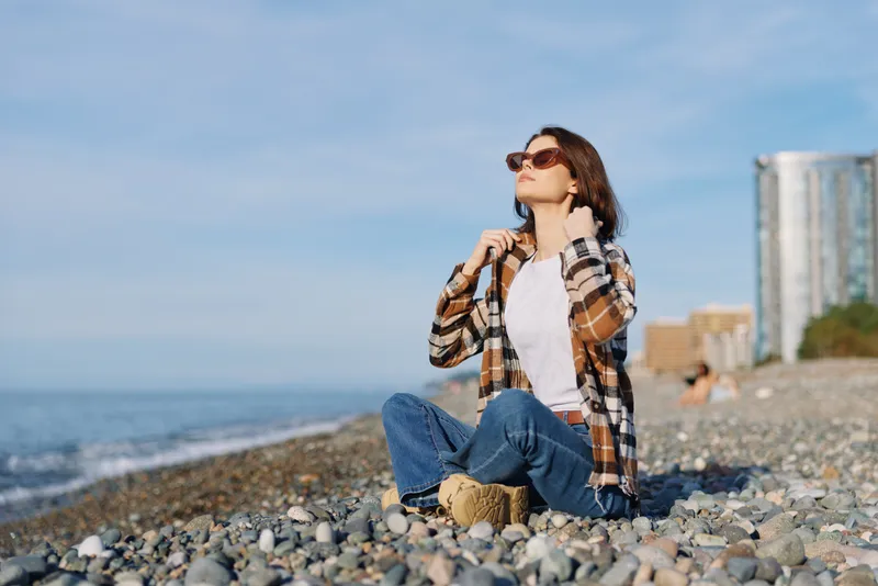 Woman sits on a pebble beach by the sea in a plaid shirt, jeans and sunglasses, enjoying a sunny day with calm waves and distant buildings on the horizon.