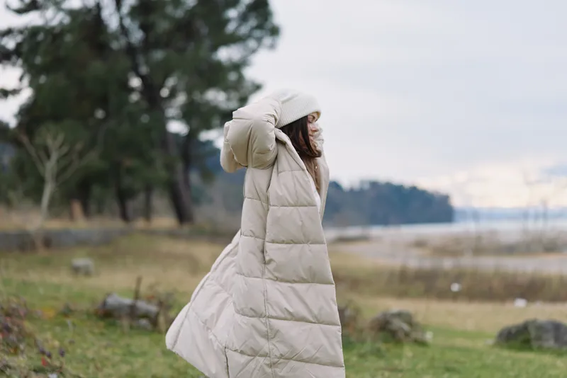 Person wearing a long beige puffer coat stands outdoors by a lake, wind lifting the garment, calm landscape with trees and shoreline, seasonal clothing, travel lifestyle, solitude and nature scene