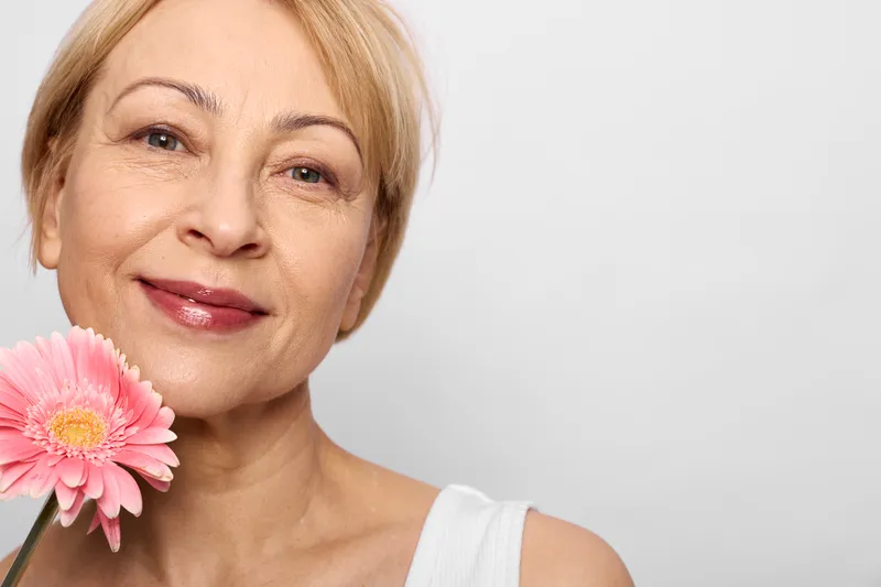Smiling mature woman portrait holding pink gerbera flower closeup on white background soft natural light beauty and happiness concept relaxed face skincare copy space