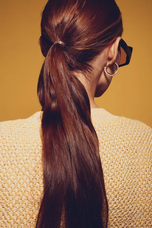 Long brown hair tied in a ponytail on a woman wearing a knitted sweater and hoop earrings. The image shows the back of her head against a warm yellow background, highlighting hairstyle and fashion.