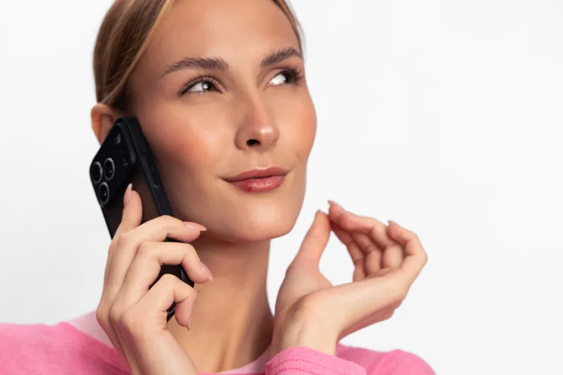 Young woman in pink sweater talking on smartphone with confident expression. Isolated on white background. Concept of communication, technology, and modern lifestyle.