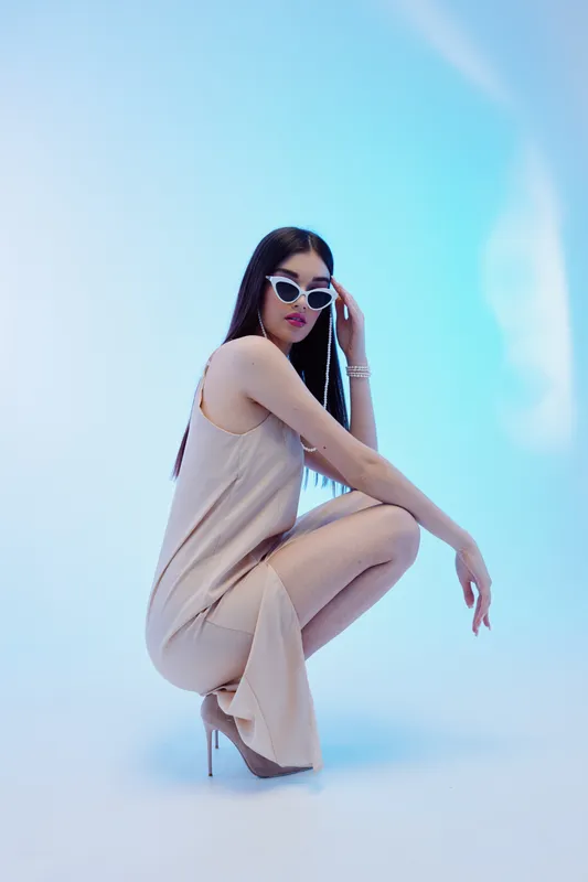 Fashionable woman with long dark hair wearing sunglasses and a beige dress squats on a blue background. Concept of beauty, style, and modern fashion trends in studio lighting.