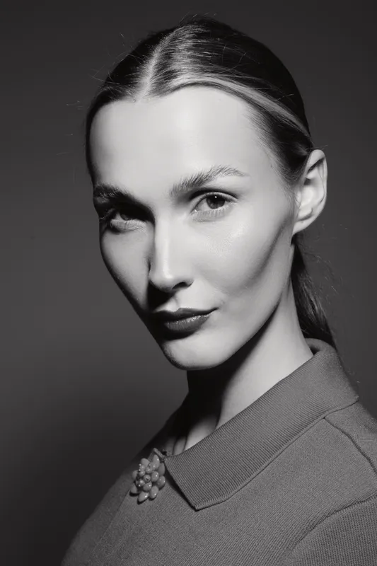 Portrait of a fair skinned Caucasian woman with slicked back hair wearing a collared garment in black and white studio lighting.