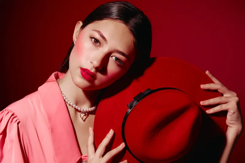 Fashionable young woman with bright makeup and glossy red lipstick holds red hat wearing soft pink blouse and pearl necklace with pendant on red background.