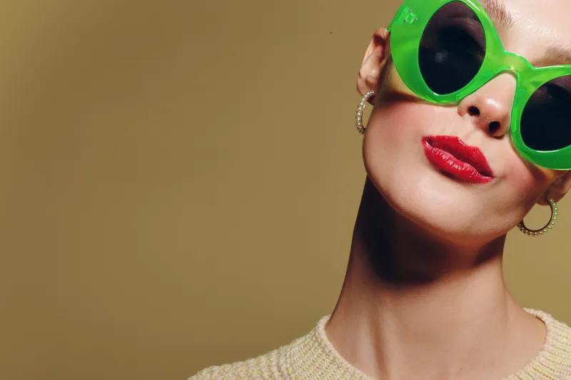 Close-up of confident woman wearing bright green sunglasses and red lipstick against beige background. Concept of fashion, style, beauty, and modern accessories.