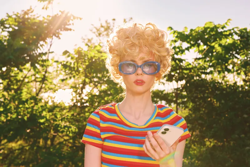 Young woman with curly blonde hair wearing a colorful striped shirt and oversized sunglasses stands outdoors in bright sun, looking at a smartphone with a surprised expression and a relaxed smile in