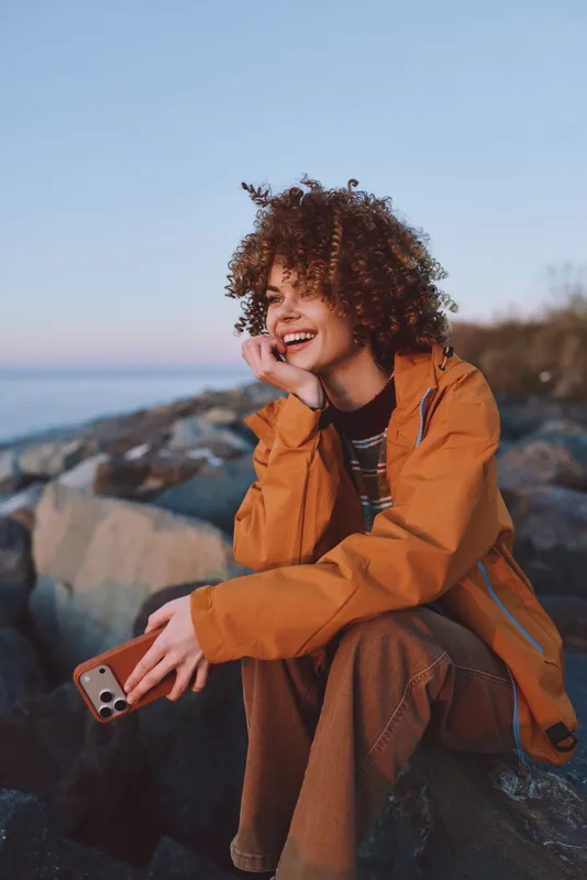 Woman lifestyle outdoors at sunset, smiling with curly hair, orange jacket, holding smartphone, coastal rocks and sea landscape, warm sky, relaxed portrait for travel, leisure
