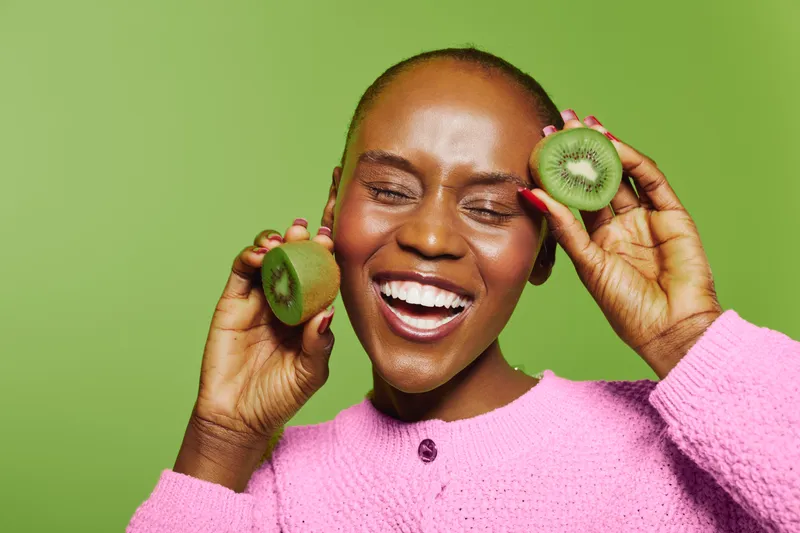 Happy dark skinned African woman with bald head smiles and holds fresh kiwi fruit slices near face. Bright green background and pink sweater create vibrant cheerful mood and healthy lifestyle concept.