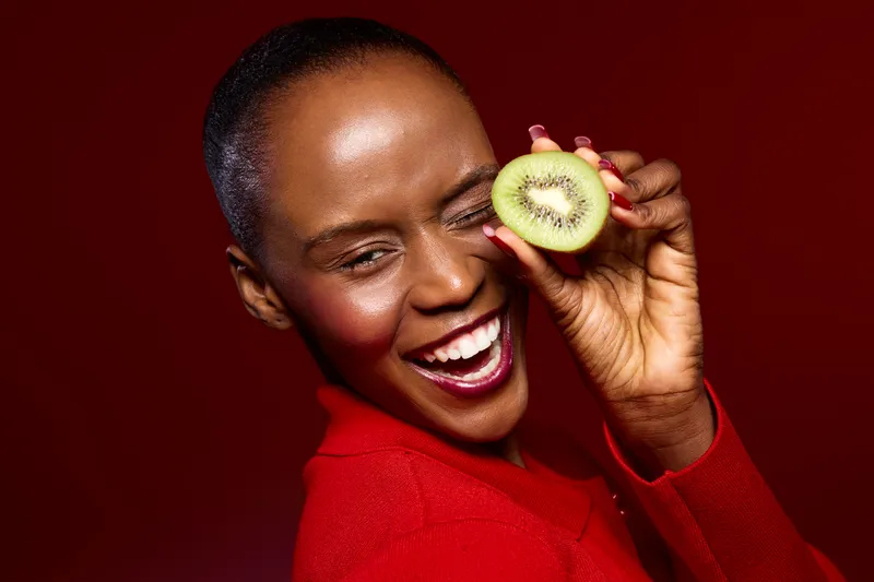 Joyful black woman with dark skin wearing red sweater smiles holding half of kiwi fruit close to her eye. Studio portrait on red background. Concept of healthy lifestyle and natural beauty.