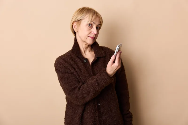 Elderly woman portrait holding a smartphone with stress worry facial expression, holding device near face against plain beige background, technology concern