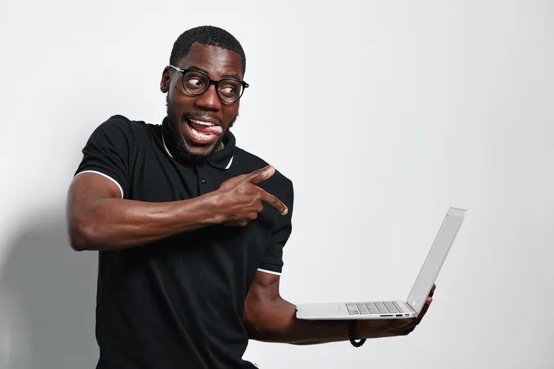 African American man with dark skin and glasses holding laptop and pointing finger with excited expression on white background in casual black polo shirt.