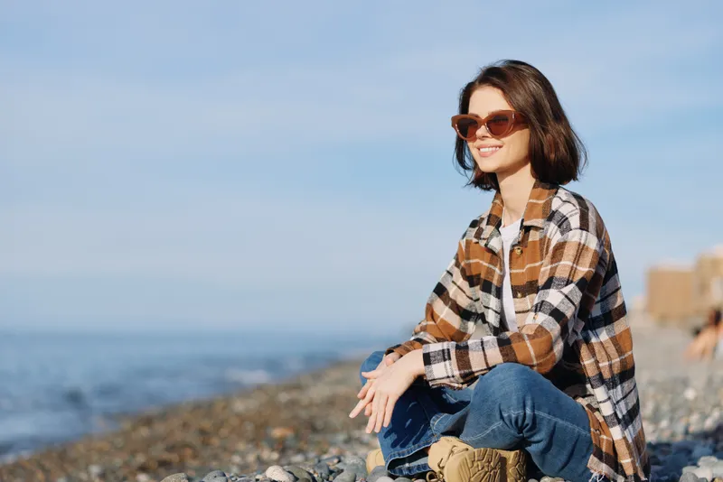 Woman in plaid shirt and jeans sits on a rocky beach with sunglasses, enjoying sunny outdoor leisure by the sea, calm horizon and sea breeze and warm light.