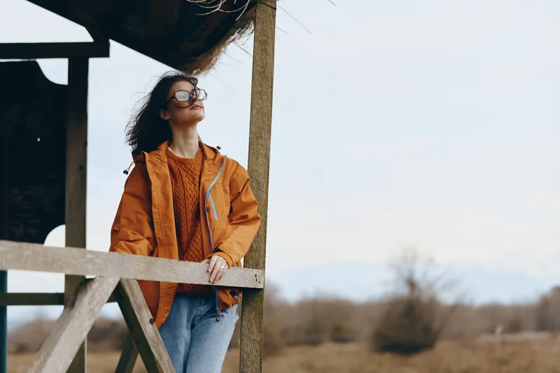 Woman lifestyle outdoor portrait, standing by wooden boards, wearing orange jacket and denim, glasses, looking to distance in warm sunlight, airy feeling, casual day outside today