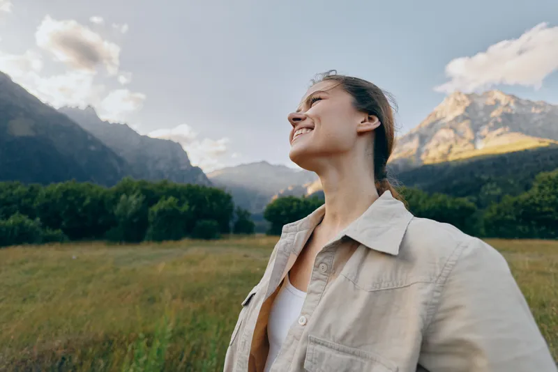 Woman enjoying outdoor lifestyle in a mountain valley, smiling with eyes closed, casual clothing and jacket, sunny day, natural scenery, carefree mood