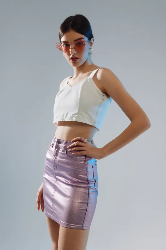 Young woman in a white crop top and shiny pink skirt poses confidently against a neutral background. Concept of woman beauty fashion and modern style in studio lighting.
