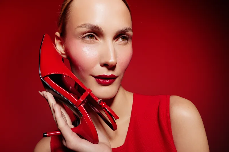 Close-up of a woman with red lipstick holding a red high heel shoe near her face against a red background. Concept of fashion, beauty, and style with vibrant colors.