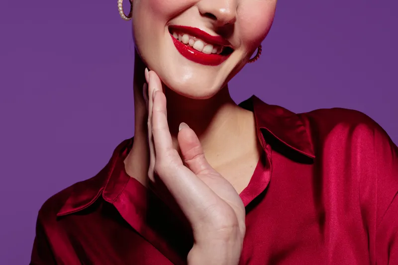 Close-up of smiling woman with red lipstick and red satin blouse touching her face on purple background. Concept of beauty, fashion, makeup, and confident female portrait.