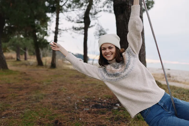 Woman on a swing outdoors, smiling in a cozy knit sweater and beanie, surrounded by trees and open space, conveying carefree leisure and natural candid energy in a serene park setting.