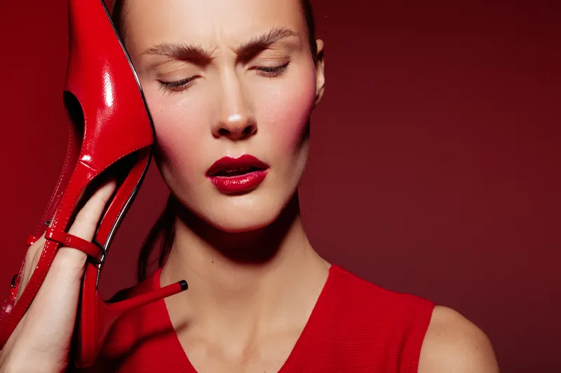 Woman in red dress holds red high heel shoe to her ear with closed eyes, expressing discomfort or pain. Red background and makeup create a bold, dramatic fashion concept.