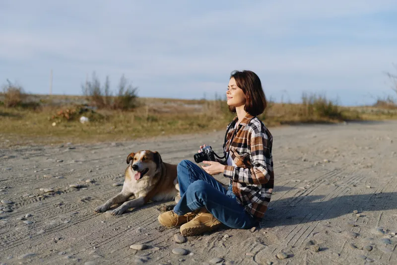 Woman sitting on sandy ground with a dog beside, holding a camera, enjoying outdoor scenery and calm breeze. Casual plaid shirt, jeans, boots capture serene nature moment.