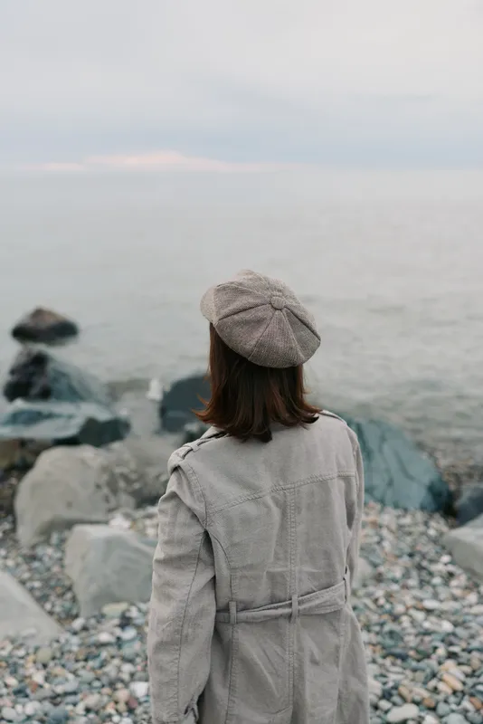 Woman in patterned beret and textured coat stands on rocky shore facing calm sea under overcast sky. Concept of autumn fashion, solitude, and peaceful coastal scenery.