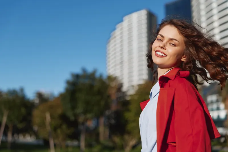 A smiling young woman in a red coat walks outdoors in an urban park with a city skyline, capturing a confident, carefree moment on a sunny day.