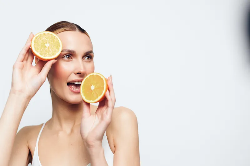 A smiling woman holds two halves of a fresh orange near her face against a white background. The image represents healthy lifestyle, freshness, and natural beauty concepts.