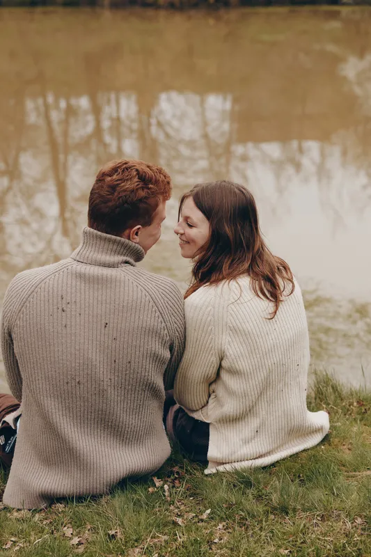 A couple sits close on a grassy bank by a calm lake, wrapped in chunky sweaters, sharing a quiet moment and a smile. A lifestyle scene portraying romance, connection, and outdoor serenity.