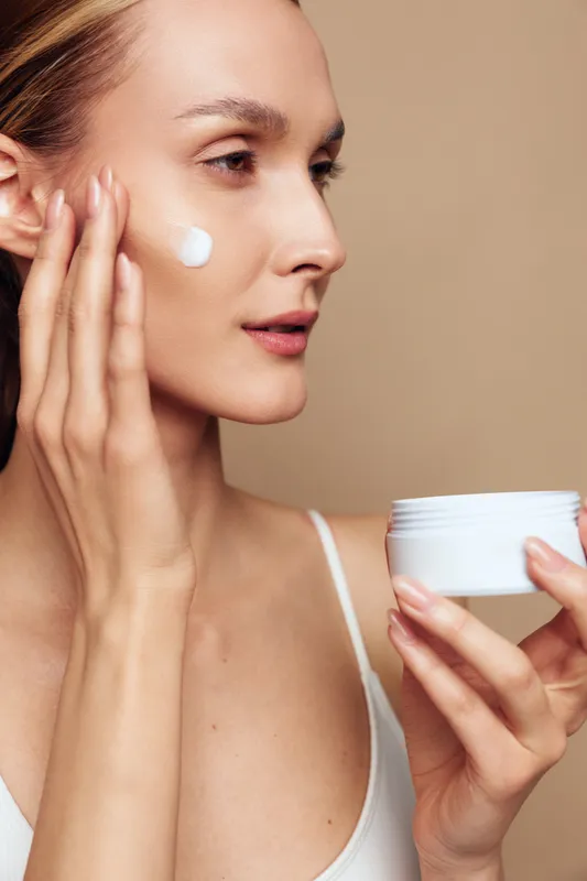 Woman applying face cream for skincare, close up beauty portrait, holding jar near cheek, indoor natural light, calm morning routine, beige background, smooth texture soft