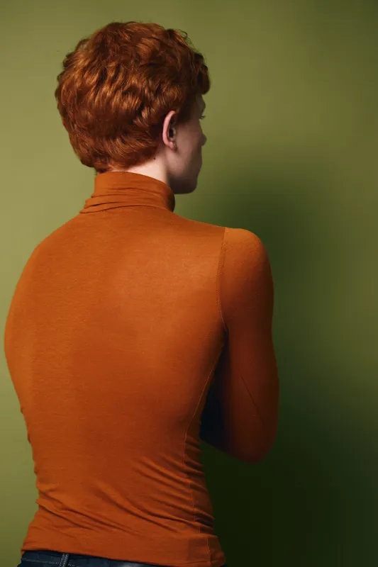 Person with red curly hair wearing orange turtleneck sweater, posing with back turned against green background in studio lighting showing texture of fabric and hair.