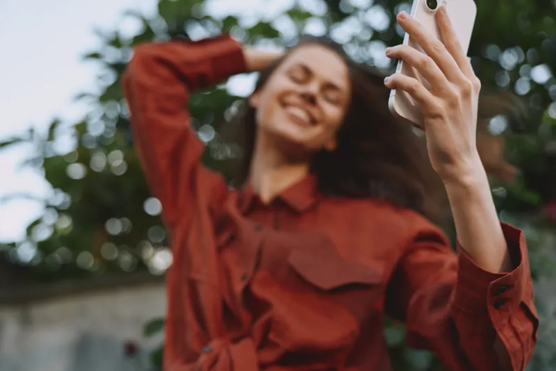 Candid young woman outdoors smiles while taking a selfie in a rust red shirt, sunlit moment, casual pose, natural light, greenery behind, lifestyle vibe