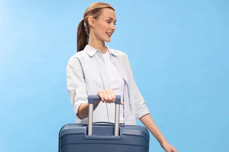 Young woman in casual clothes smiling while holding a suitcase handle against blue background. Concept of travel, vacation, tourism, and summer holiday preparation.