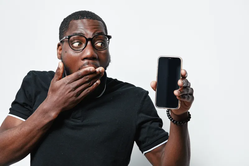 African American man with dark skin and glasses showing smartphone with blank screen and covering mouth in surprise or shock on white background.