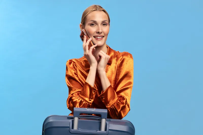 Smiling woman in orange silk blouse talking on phone with suitcase in front. Blue background studio portrait. Concept for travel, communication, business, and lifestyle.