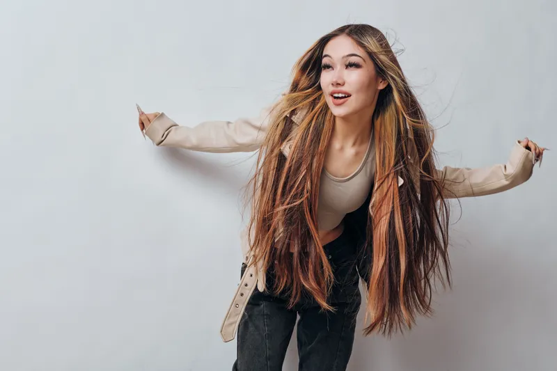 Young woman with long braided hair wearing beige jacket and black pants posing happily with arms outstretched on white background in casual style fashion portrait.