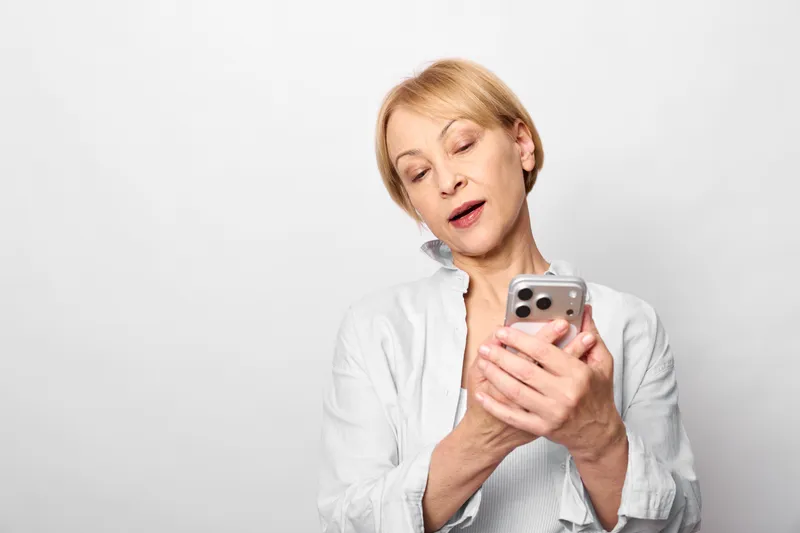 Woman with smartphone looking surprised, eyes closed, holding phone near face, indoor portrait with emotional moment, soft light, casual shirt, copyspace for text