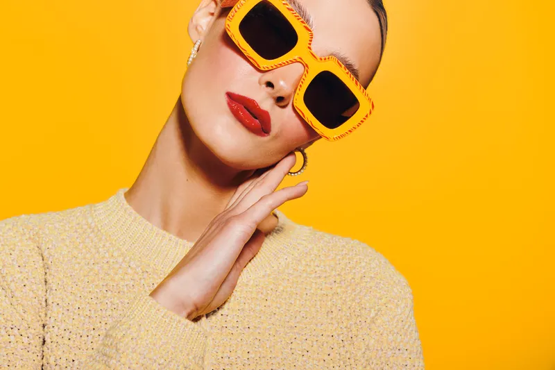Stylish woman with bright orange sunglasses and red lipstick poses against yellow background wearing beige sweater. Concept of fashion, summer style, and confident attitude.