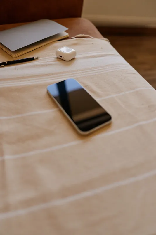 A cozy home scene shows a smartphone on a striped bedspread beside a notebook and wireless earbuds case, capturing a calm, organized lifestyle moment for everyday routine.
