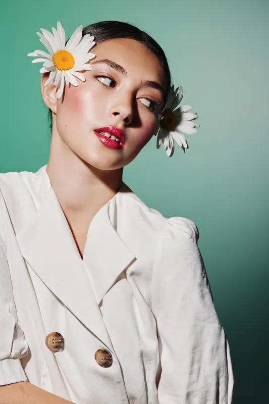 Stylish young woman with flowers in hair poses confidently in white outfit against green backdrop. Glossy makeup and creative beauty look emphasize modern fashion aesthetics.
