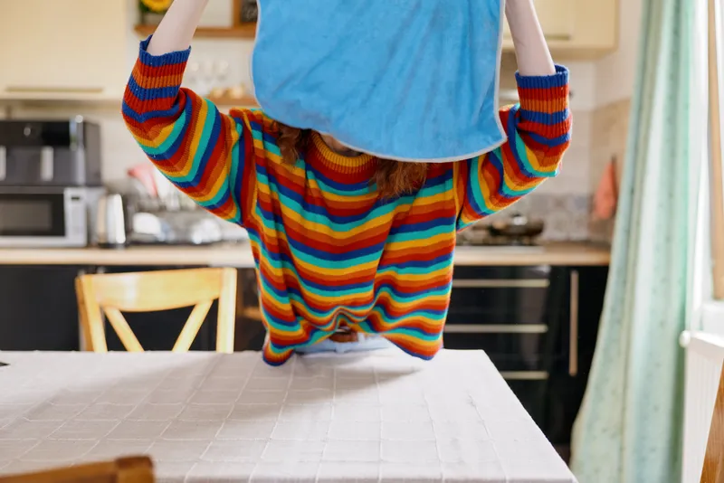 A young child in a rainbow striped sweater hides under a blue cloth in a bright kitchen, turning a simple moment into playful imagination at home.