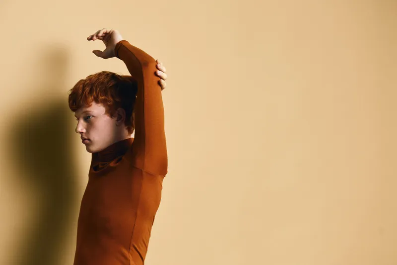 Young man with red hair wearing brown fitted shirt stretching arm against beige wall in minimal indoor environment. Profile portrait with soft shadow and natural lighting.