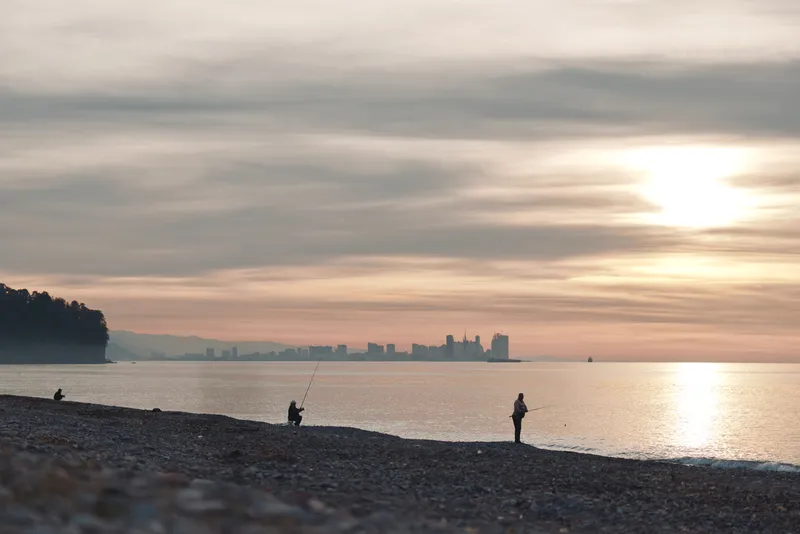 Beach at sunset with calm water and silhouettes walking along the shore, distant city skyline on the horizon, creating a tranquil coastal scene of leisure and nature.