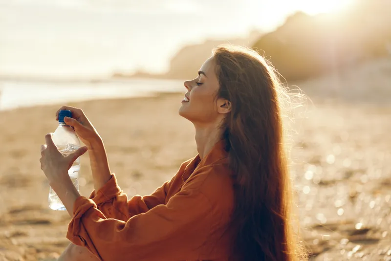Young woman with long brown hair enjoys a sunny day sitting on the beach while holding a water bottle and smiling peacefully with closed eyes.