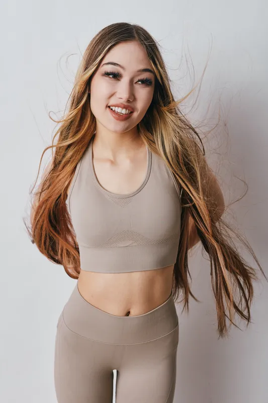 Young woman with long hair wearing beige workout clothes smiles standing against white background. Fitness model, healthy lifestyle, sport apparel and casual activewear for exercise or yoga.
