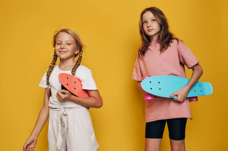 Two young girls stand against a yellow background holding colorful skateboards. They wear casual summer clothes and express confidence and joy.