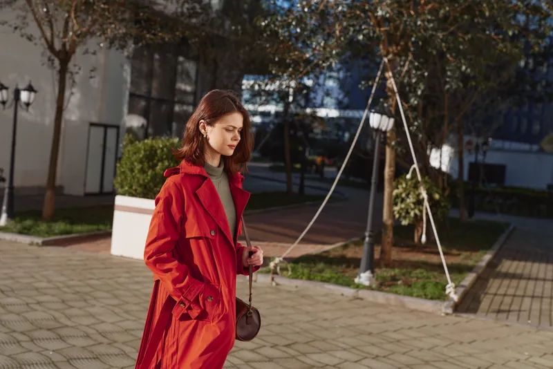 Young woman wearing a bright red trench walks through a sunlit urban square, her calm expression and steady pace conveying quiet confidence.