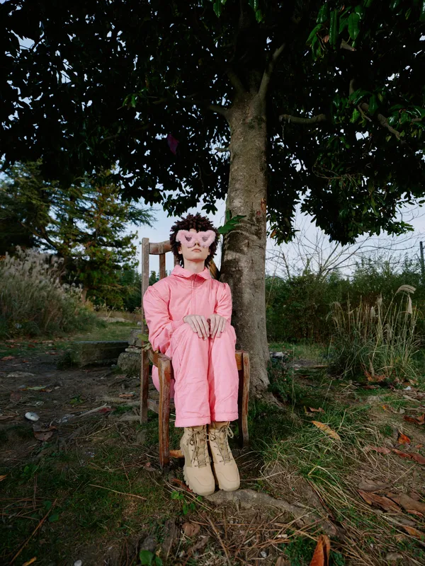 Doll seated under a tree in an outdoor garden wearing a pink coat and tall boots, still pose with natural backdrop and tranquil mood for playful lifestyle imagery